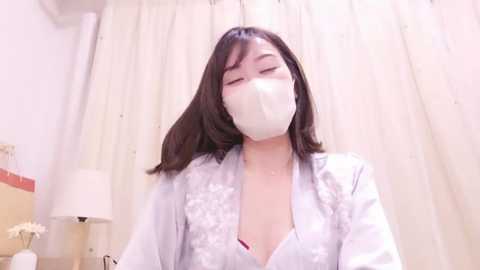 Media: Video of an Asian woman with straight black hair, wearing a white satin robe with floral embroidery and a white face mask, sitting in a dimly lit room with white curtains and a beige lamp.