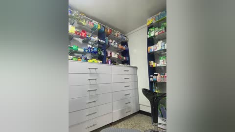 Media: Video of a modern pharmacy interior featuring white cabinets filled with various pharmaceutical products. Brightly colored medications and health supplies are neatly organized on shelves. A black chair sits in the corner.