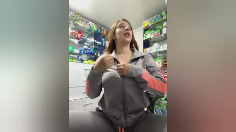 Media: Video of a young woman with light skin and long brown hair in a gray zip-up hoodie, standing in a brightly lit pharmacy aisle, surrounded by colorful medicine shelves.