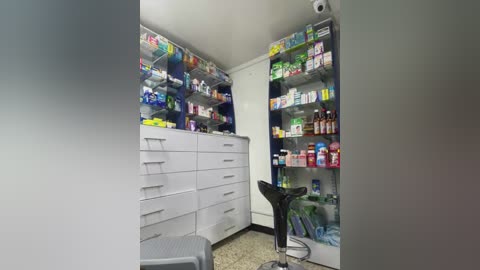 Media: Video of a small, well-organized pharmacy with white cabinets filled with colorful medicines, a black stool, and a counter with a glass jar.