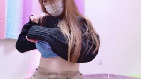 Media: A video of a young woman with long, straight, light brown hair, wearing a black sweater, blue lace bra, beige pants, and a face mask, standing in a brightly lit room with a pink and blue striped background.