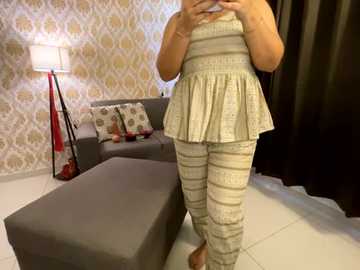 Media: Video of a light-skinned woman in a striped, sleeveless top and patterned pants, standing in a modern living room with gray furniture, white wallpaper, and a black curtain.