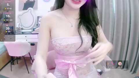 Media: Video of an Asian woman with long black hair, wearing a strapless pink lace dress with a bow, in a bedroom with white furniture, pink curtains, and a chair.