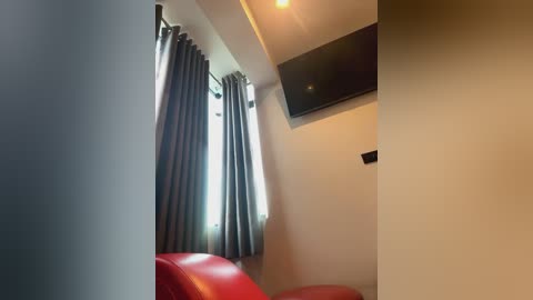 Media: Video of a modern, minimalist bedroom with beige walls, a large window with dark grey curtains, a red leather chair, and a mounted flat-screen TV.