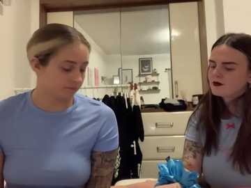 Media: Video of two young women in a bedroom, one with blonde hair, the other with long brown hair, wearing light blue shirts, discussing tattoos on their arms.