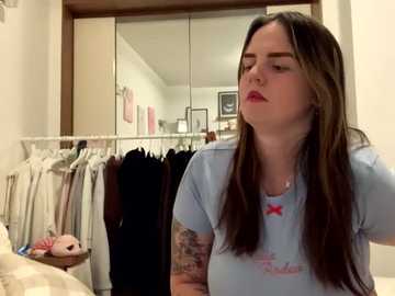 Media: Video of a young woman with long brown hair, wearing a light blue T-shirt with a pink bow, standing in a cluttered bedroom with a white wardrobe and hanging clothes.