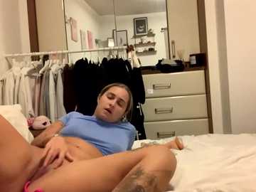 Media: Video of a light-skinned woman with brown hair, wearing a blue crop top, lying on a bed, masturbating, in a bedroom with a mirror, white dresser, and hanging clothes.