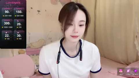 Media: Video of a young East Asian woman with light skin, brown hair in a bun, wearing a white T-shirt, seated in a pastel bedroom with a pink bedspread, displaying a smartphone screen with weather and temperature information.