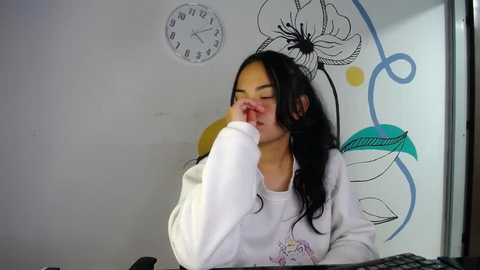 Media: Video of a young woman with long black hair, wearing a white sweatshirt, covering her mouth with her hand, in a room with a white wall and a large, colorful floral design.