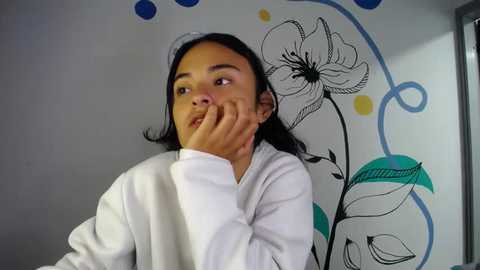 Media: Video of a young girl with medium brown skin, wearing a white robe, covering her face with her hand. Background features a colorful, abstract floral mural with blue, yellow, and green accents.