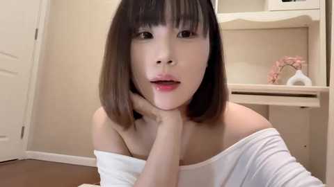 Media: Video of an East Asian woman with straight, shoulder-length black hair, wearing a white off-shoulder top, resting her chin on her hand, in a minimalist, beige-toned room with a wooden desk and a closed white door.