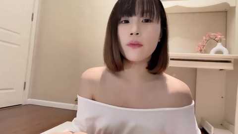 Media: A video of an Asian woman with a straight bob haircut, wearing an off-the-shoulder white top, sitting indoors on a wooden floor. Background includes a beige shelf with pink flowers, a white door, and a white desk.