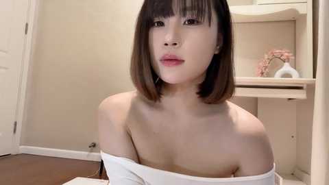 Media: A video of an Asian woman with shoulder-length straight black hair and pale skin, wearing a white off-shoulder top, sitting indoors with beige walls and a wooden floor.