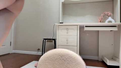 Media: A video of a minimalist white desk with drawers, a black stool, and a pink vase, in a clean, beige-walled room with wooden flooring.