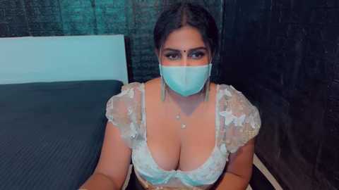 Media: A video of a South Asian woman with medium brown skin, dark hair in braids, wearing a light blue mask and revealing sheer white lace lingerie with puffed sleeves. She sits on a black bed with a white wall behind her.