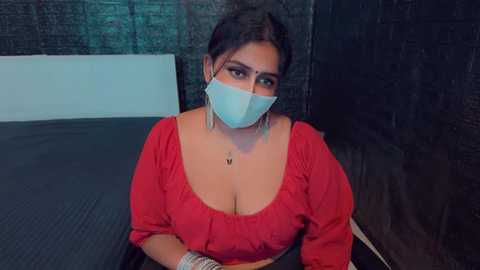 Media: Video of a South Asian woman with medium-dark skin tone and dark hair in a ponytail, wearing a red blouse, face mask, and dangling earrings, sitting on a dark surface, with a brick wall background.