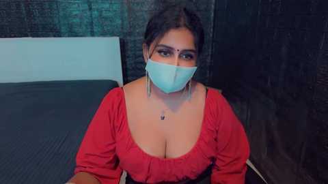 Media: Video of a South Asian woman with medium brown skin, dark hair, wearing a red blouse with a plunging neckline, a face mask, and a bindi. She sits on a black mat in a dimly lit, brick-walled room.