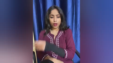 Media: Video of a South Asian woman with medium brown skin, long black hair, and a maroon embroidered blouse, sitting on a black chair against a blue backdrop, looking thoughtful.