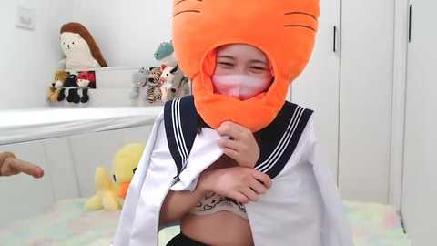 Media: Video of a young girl in a bright orange cat mask, white sailor outfit, and pastel-colored room with stuffed animals and toys.