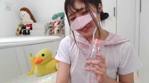 Media: Video of an Asian woman with light skin, dark hair in a ponytail, wearing a white T-shirt, pink mask, and holding a pink straw, in a white bathroom with a duck toy and stuffed animals.