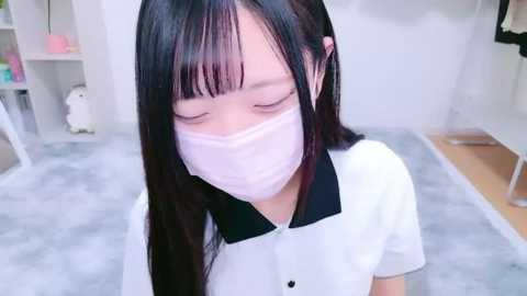 Media: Video of an East Asian woman with long black hair, wearing a white blouse and pink face mask, sitting on a gray rug in a minimalist, light-colored room with shelves and a white wall.