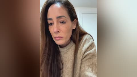 Media: A close-up video of a woman with long, straight brown hair, wearing a beige sweater, looking introspective against a blurred, light-colored background.