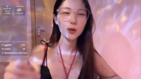 Media: Video of an East Asian woman with long dark hair, wearing glasses, a black top, and multiple necklaces, blowing a kiss, surrounded by a pinkish glow.