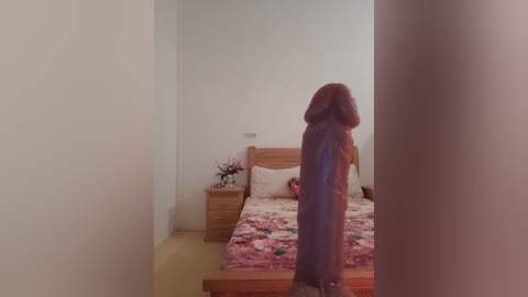 Media: A video of a small, round, fluffy dog standing on a bed with a floral-patterned blanket, against a plain white wall, in a simple, minimalistic bedroom.