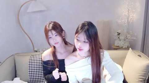 Media: Video of two East Asian women with long hair, one in a black houndstooth dress, the other in a white sweater, sitting on a beige couch in a minimalist, softly lit living room.