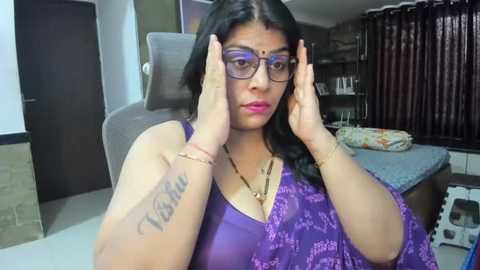 Media: Video of a South Asian woman with medium skin tone, wearing purple glasses, adjusting her glasses, in a modern living room with a gray couch and brown curtains.