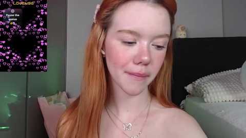 Media: Video of a fair-skinned, red-haired young woman with freckles, wearing a simple necklace, in a cozy bedroom with a TV displaying a game in the background.