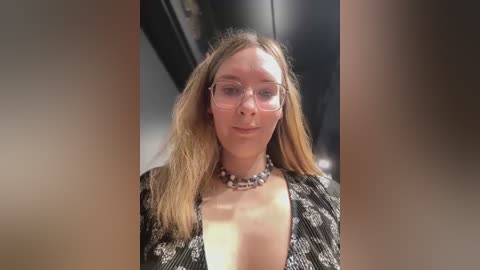 Media: A video of a fair-skinned woman with long, blonde hair, wearing large, clear glasses and a black sequined dress, set against a blurred background, featuring a necklace with a floral pattern.