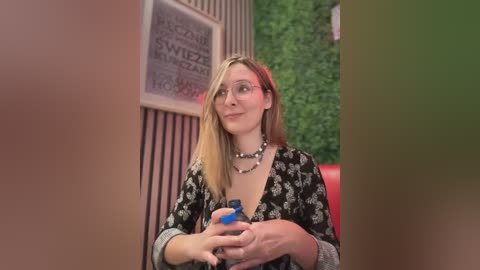 Media: Video of a blonde woman with glasses and a floral dress, holding a blue bottle, standing in a room with green wall art and a red chair.