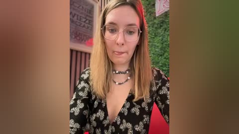 Media: Video of a young woman with light skin, shoulder-length blonde hair, wearing glasses and a black floral-patterned dress, seated in a cozy indoor setting with red lighting and greenery.