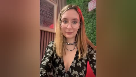 Media: Video of a young Caucasian woman with straight blonde hair, wearing glasses, a black floral dress, and multiple necklaces, standing in front of a green wall with red and black decor.