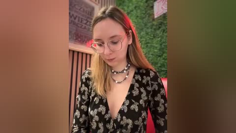 Media: Video of a young Caucasian woman with light skin, blonde hair, and glasses, wearing a black floral dress with a deep V-neckline and a black and white beaded necklace, standing indoors with green foliage and red lighting in the background.