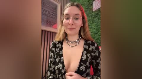 Media: Video of a Caucasian woman with long, straight blonde hair, wearing large glasses and a black floral dress with a deep V-neckline, standing in front of a green wall and a metal fence.