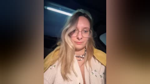 Media: Video of a young woman with long blonde hair, wearing glasses, a yellow and white floral-patterned blouse, and a silver necklace, seated in a dimly lit car.