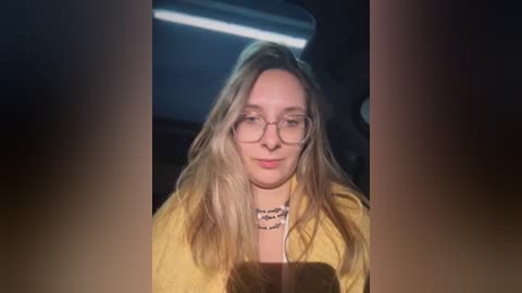 Media: Video of a blonde woman with glasses, wearing a yellow jacket, and a black choker necklace, sitting in a dimly lit car with a blue overhead light.