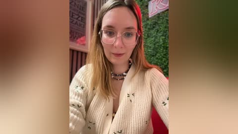 Media: Video of a fair-skinned, blonde woman with glasses and a cream-colored sweater, seated indoors with a red cushioned background, wearing a beaded necklace.