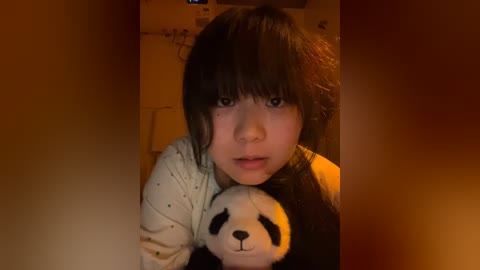 Media: Video of a young Asian girl with freckles, black hair, and a white shirt, holding a stuffed panda, in a dimly lit room with a warm, orange glow.