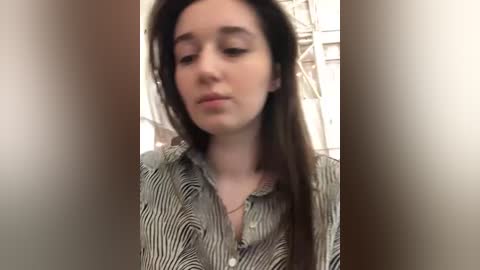 Media: Video of a young woman with straight, dark hair, wearing a striped shirt, looking downward with a neutral expression. Background is blurred, featuring a white wall with a pattern.