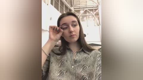 Media: Video of a young woman with straight brown hair, wearing a black-and-white striped shirt, adjusting her glasses indoors with industrial background.