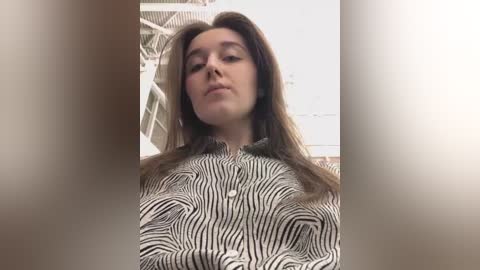 Media: Video of a young woman with long brown hair, wearing a striped button-up shirt, standing in a modern, minimalist room with white walls and a metal staircase.