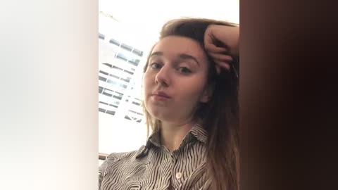 Media: Video of a young Caucasian woman with fair skin and straight brown hair, wearing a striped button-up shirt, resting her head on her hand, in a dimly lit room with vertical blinds.