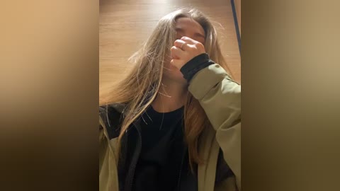 Media: Video of a young woman with long brown hair, wearing a black top and olive-green jacket, covering her face with one hand, in a dimly lit hallway with wooden flooring.