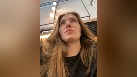 Media: Video of a fair-skinned woman with long, straight brown hair, wearing a black top, sitting in a salon chair under bright overhead lights.