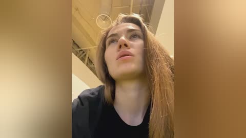 Media: Video of a Caucasian woman with light skin, long brown hair, and blue eyes, wearing a black shirt, taken in an indoor setting with a beige wall and exposed ceiling ducts.