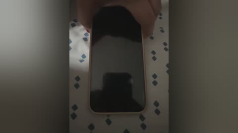 Media: A video showing a hand holding a rectangular smartphone with a black screen, reflecting a person\u2019s shadow on a tiled wall. The background is dimly lit, with a beige wall and a patterned white tile floor.