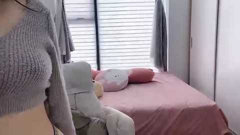 Media: Video of a woman in a gray knit sweater, standing near a pink bed with white pillows, in a minimalist, well-lit room with white blinds and a wardrobe.